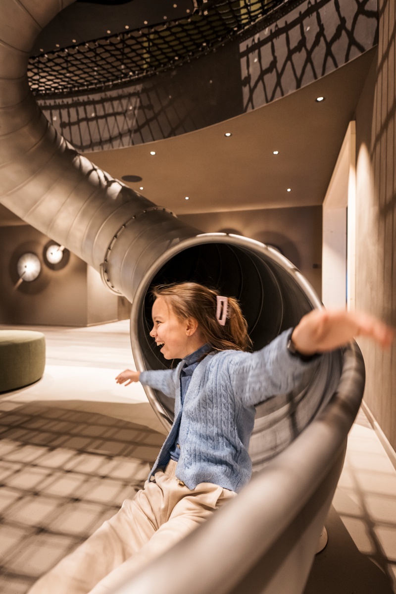 Ein lachendes Mädchen kommt aus einer Tunnelrutsche in einem Indoor Spielplatz, ihre Arme sind zur Seite ausgestreckt © Alex Filz 