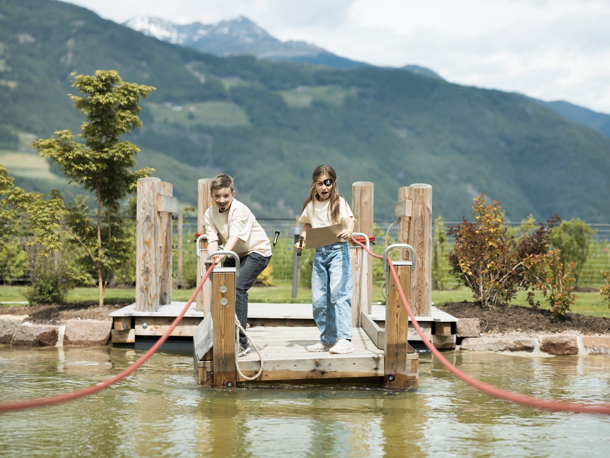 Two children stand on a rope ferry in the water, with trees and mountains visible in the background © Alex Filz