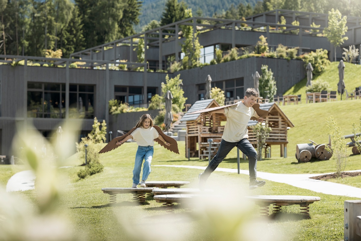 Two children with wings on their backs are jumping on the jumping discs. Buildings, trees and other wooden playground equipment can be seen in the background. The weather is sunny © Alex Filz