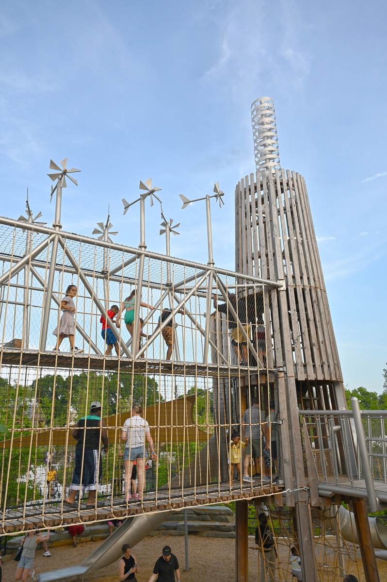 Children climb a multi-storey wooden tower via a two-storey bridge. The bridge is enclosed by ropes and nets. There are several wind turbines on the bridge piers and a double helix on the wooden tower.