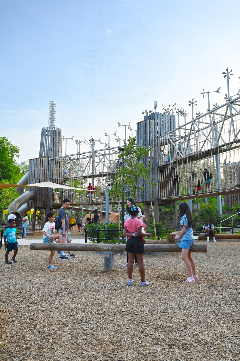 Children are playing on a wooden seesaw. In the background are multi-storey wooden towers connected by a two-storey bridge. There is a double helix on one tower and many wind turbines on the bridges.