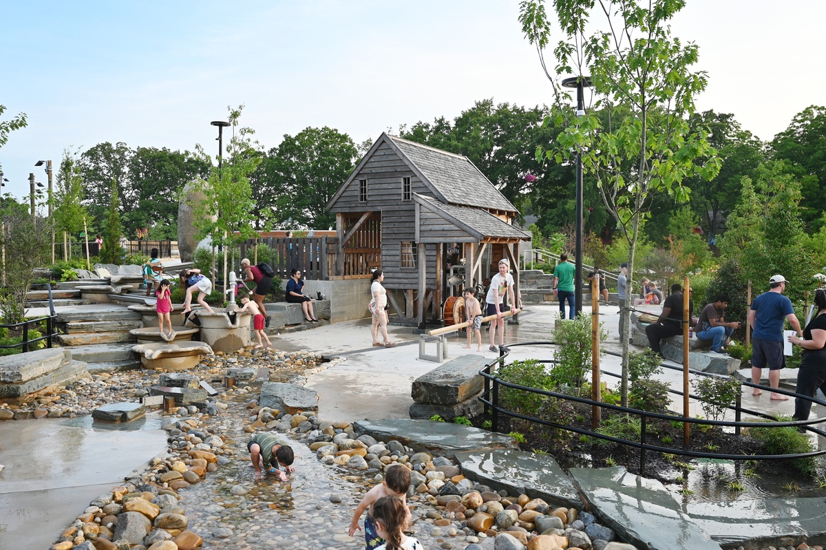 Children play and splash around in a water play area. The watercourse comes from a wooden play mill house.