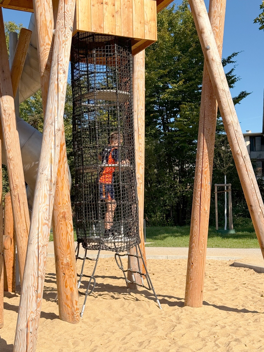 A child climbs from the bottom to the top through a black net tunnel into a wooden play tower. 