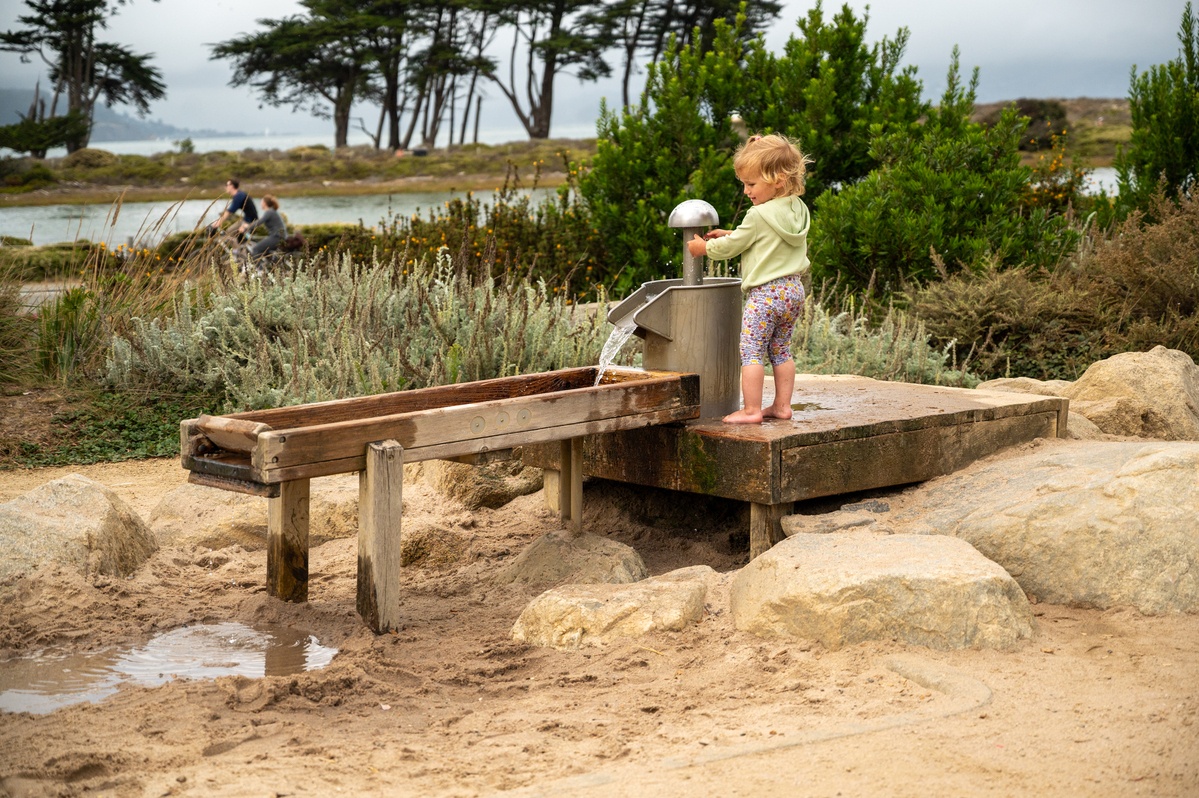 Kleinkind spielt barfuß an einer Wasser-Quelldüse, naturnaher Wasserspielplatz nahe der Golden Gate Bridge.