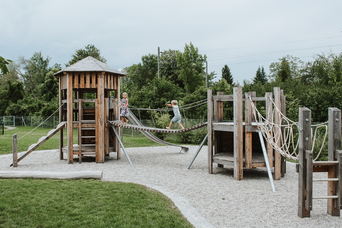 Ein naturnaher Spielplatz mit spielenden Kindern auf einer Hängebrücke zwischen großer Sechseckhütte mit Dach und Sechseckhütte.