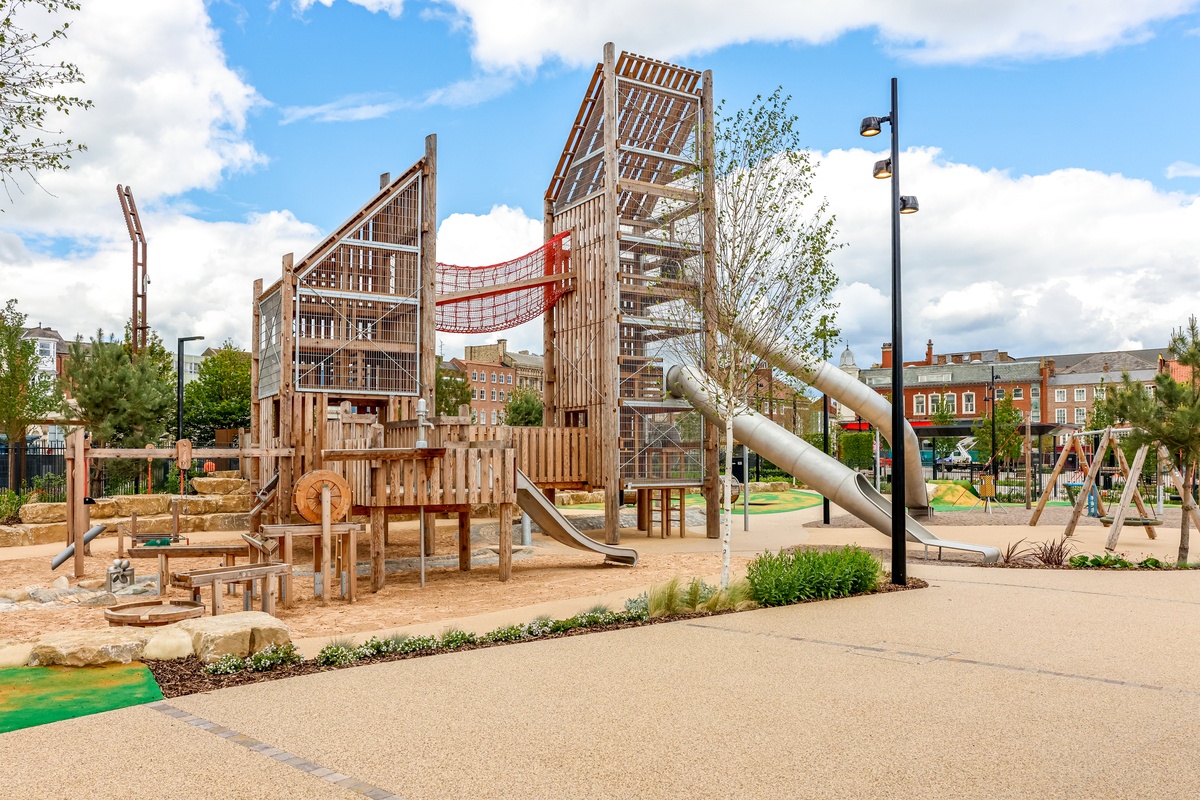 Two towers connected by a red suspension bridge with child sand and water play beneath it.  