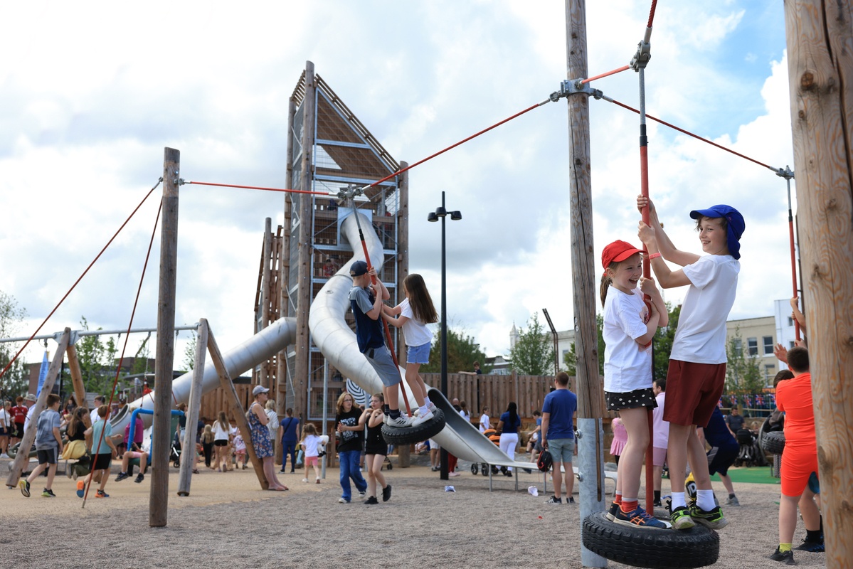 Children run across stockholm play space while four children stand laughing on the tire swings.