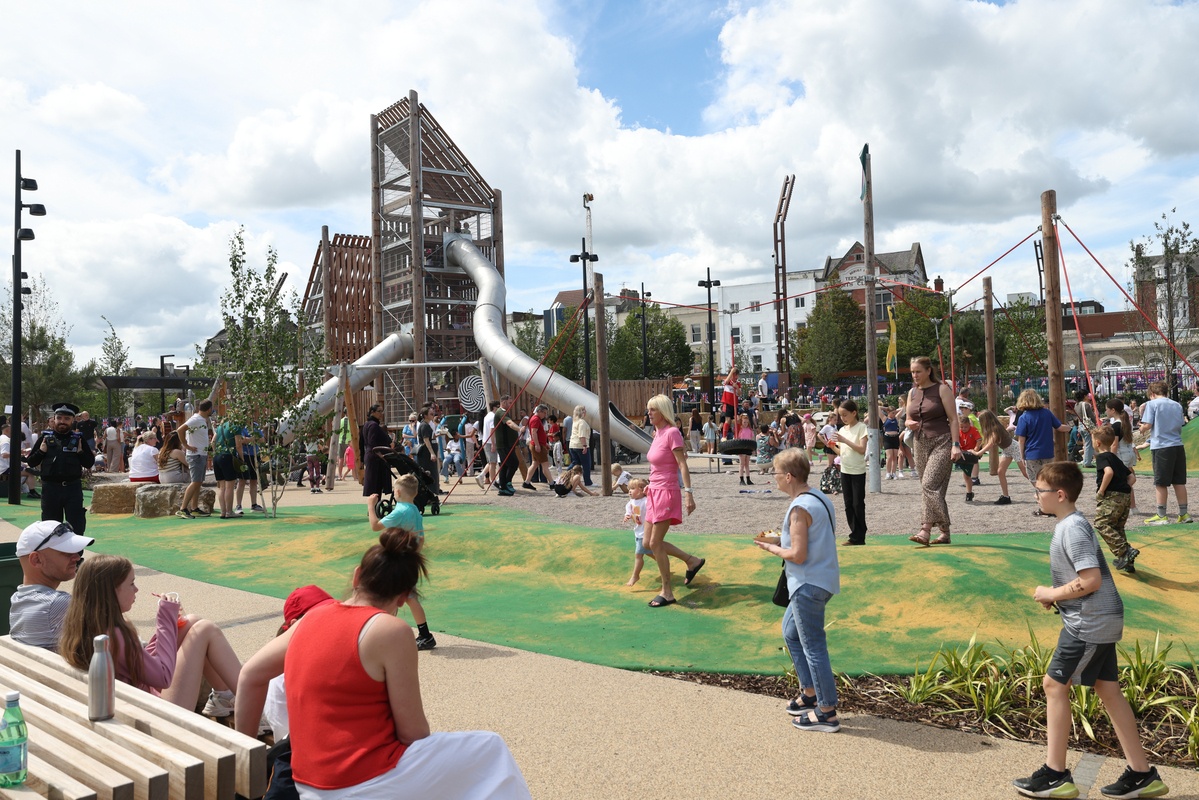 Many families crowd playspace while onlookers obseve from a bench.