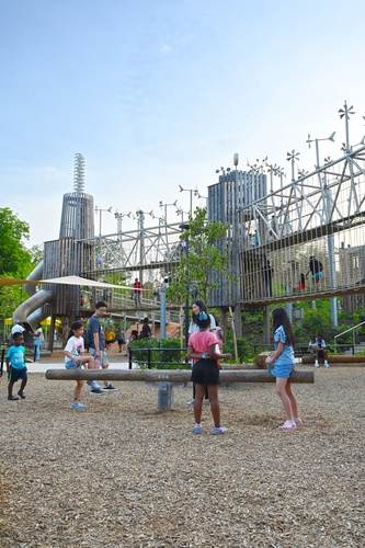Children are playing on a wooden seesaw. In the background are multi-storey wooden towers connected by a two-storey bridge. There is a double helix on one tower and many wind turbines on the bridges.