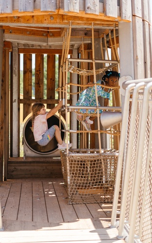 Two children are climbing on a natural-coloured net tunnel. The entrance to a tunnel slide can be seen in the background.