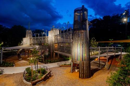 At dusk, you can see a climbing and play combination consisting of wooden towers that can be accessed via a ramp. The floors of the towers are connected by a two-storey bridge. Street lamps illuminate the area.