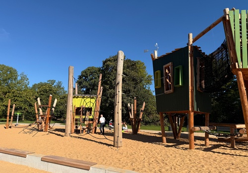 Große Kletterkombination "Skywalk" im Johannes-Giesberts-Park Köln bestehend aus verschiedenen durnkelgrünen Spielboxen und Türmen. Die Türme sind durch einen schwarzen Netztunnel miteinander verbunden. Im Hintergrund blauer Himmel, Boden aus Sand.