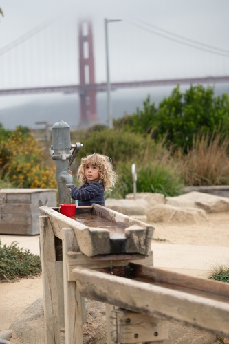 Kleinkind spielt an einer Wasserpumpe mit Holzrinne auf einem Wasserspielplatz, die Golden Gate Bridge ist im Hintergrund zu sehen.