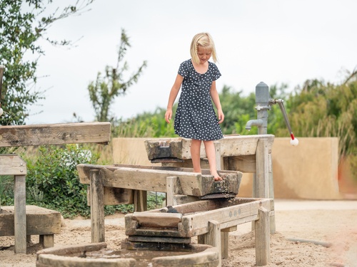 Kind balanciert über eine Wasserrinne aus Holz auf einem Abenteuerspielplatz. Im Hintergrund die Golden Gate Bridge.