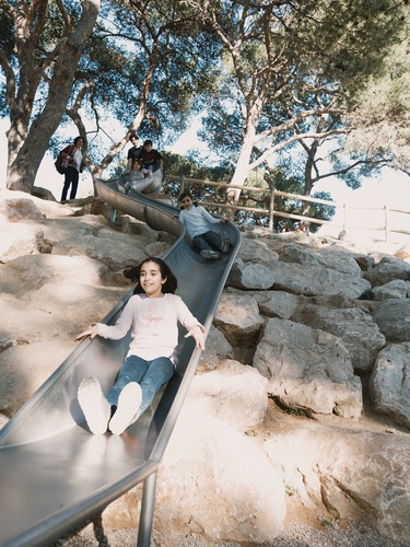Kinder rutschen eine Edelstahlrutsche hinunter. Die Rutsche befindet sich an einer großen Steinmauer.