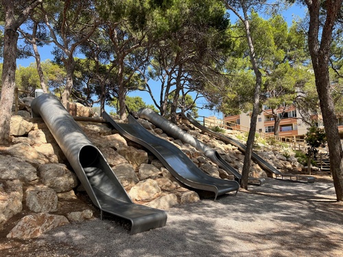 Various stainless steel slides in Castell de Bellver Park on a slope with large rocks.