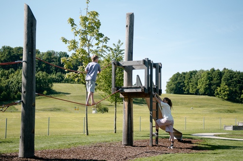 One child balances across a walk rope to a platform. Another climbs a ladder up to the platform.