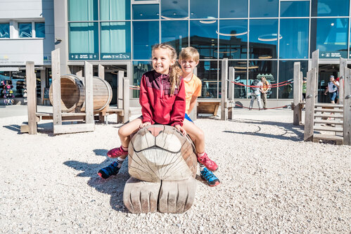 Two children sit laughing on a hand-carved wooden animal sculpture in front of the Jennerbahn mountain station. Gravel area and mountain backdrop in the background.