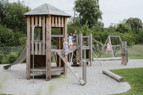 Naturnaher Spielplatz mit spielenden Kindern. Im Vordergrund ist eine Sechseckhütte mit Dach, Steigstamm und Rutschstange zu sehen.