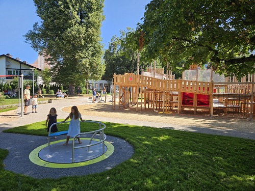 Kinder spielen auf einem Karussell vor einer großen naturnahen Holz-Spielanlage.