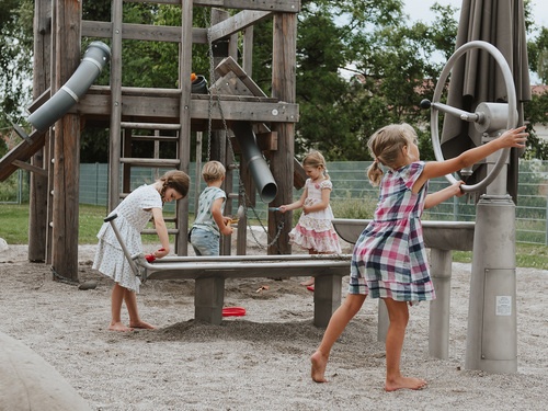 Children playing at a water flooter with crank handle pump and water basin with seal. A building site can be seen in the background.